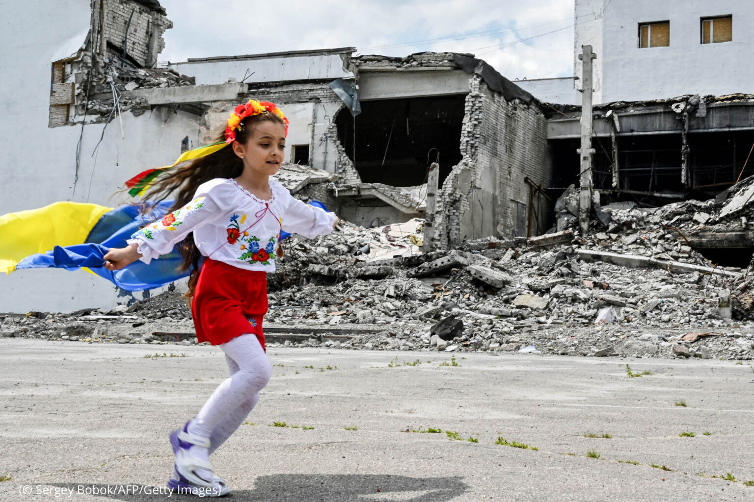 Ein Mädchen läuft mit einer ukrainischen Fahne. Im Hintergrund sind zerstörte Gebäude zu sehen. Das Foto stammt 